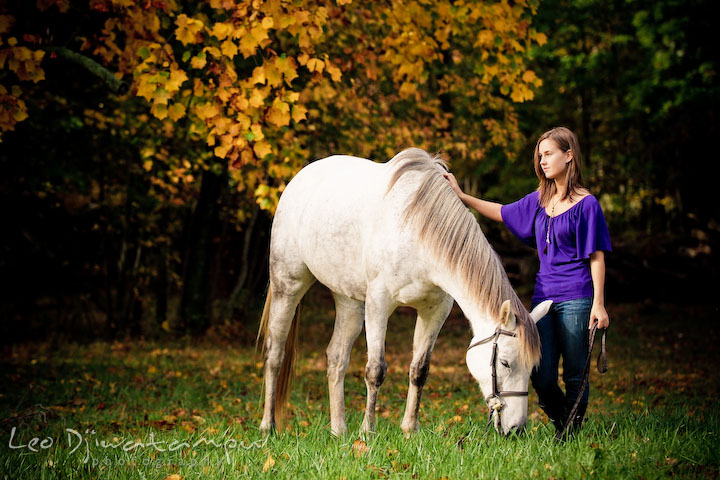 Girl standing next to horse who's eating grass. Annapolis Kent Island Maryland High School Senior Portrait Photography with Horse Pet by photographer Leo Dj