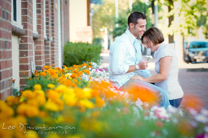 Engaged guy give flower to his fiance, sitting by flower bed. Candid Old Town Alexandria Virginia Engagement Photography Session by Wedding Photographer Leo Dj