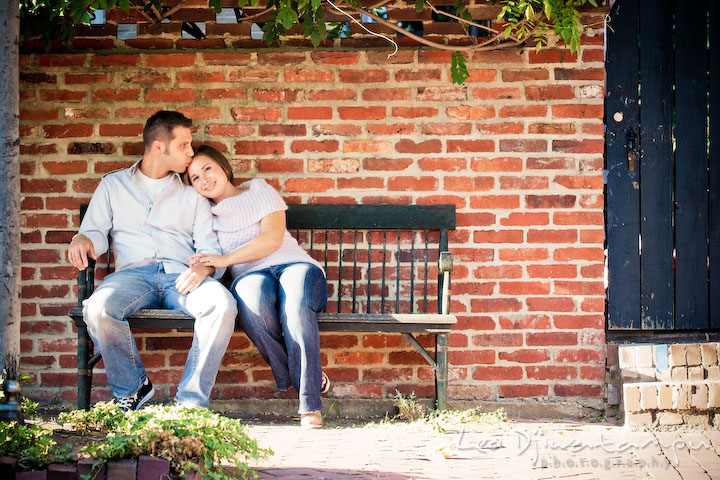 Engaged guy kissed his fiancee on a bench outside. Candid Old Town Alexandria Virginia Engagement Photography Session by Wedding Photographer Leo Dj