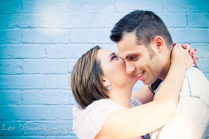 Engaged girl kissed her fiancee by a light marine blue brick wall. Candid Old Town Alexandria Virginia Engagement Photography Session by Wedding Photographer Leo Dj