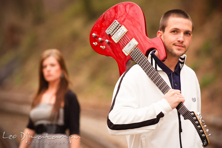 Engaged guy posing with his red electric guitar. His fiancée in the background. Pre-Wedding Engagement Photo Session at Sykesville Maryland with Train Rail and Caboose by wedding photographer Leo Dj Photography