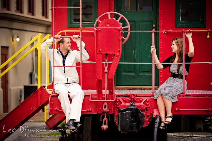 Engaged guy and his fiancée on a red caboose looking at each other and smiling. Pre-Wedding Engagement Photo Session at Sykesville Maryland with Train Rail and Caboose by wedding photographer Leo Dj Photography