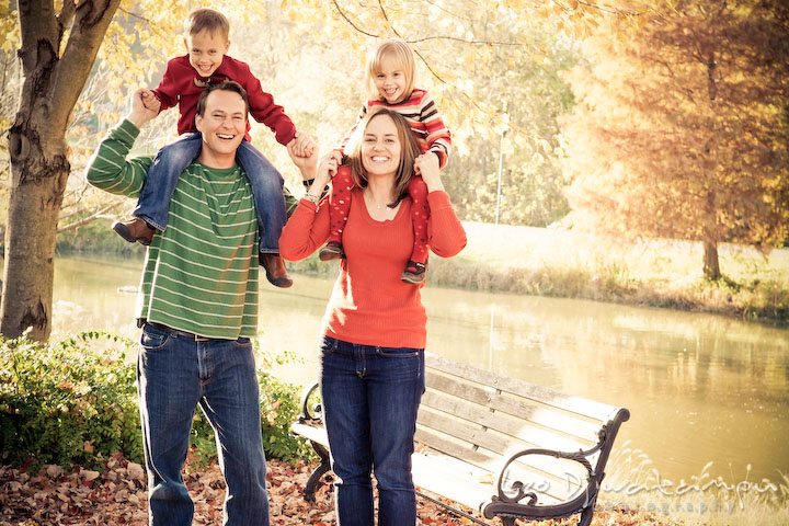 childrend on mom and dad's shoulder, laughing. pond, bench and fall colored foliage. Fun candid family children lifestyle photographer Annapolis Maryland