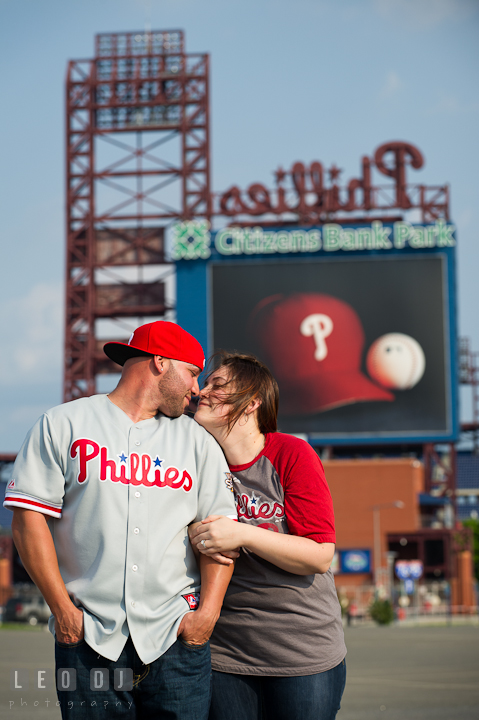 Engaged guy and his fiancée wearing Phillies clothing cuddling. Pre-wedding or engagement photo session at Phillies Ball Park, Love Park, Philadelphia, by wedding photographers of Leo Dj Photography.