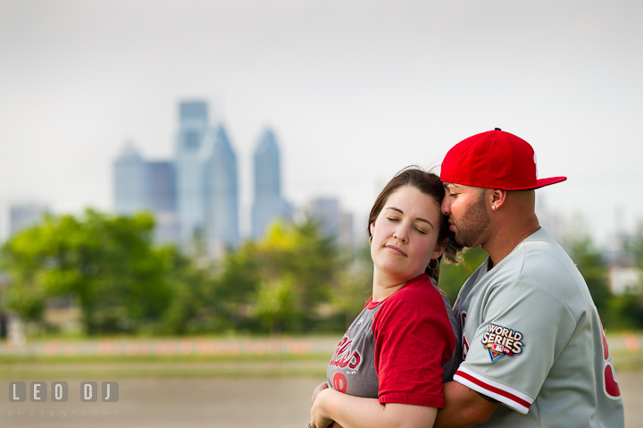 Engaged couple with Phillies merchandise cuddling, with Philadelphia skyscrapers in the background. Pre-wedding or engagement photo session at Phillies Ball Park, Love Park, Philadelphia, by wedding photographers of Leo Dj Photography.