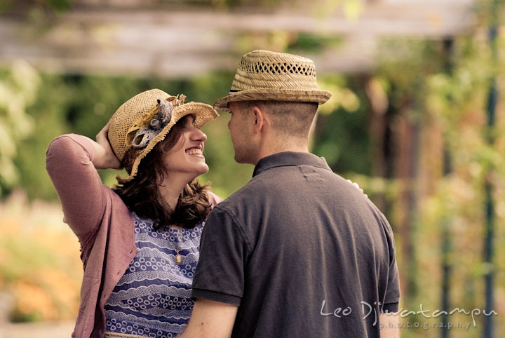 Engaged girl wearing hat and smiling big to her fiancé. Pre-wedding engagement photo session at Brookside Gardens, Wheaton, Silver Spring, Maryland, by wedding photographer Leo Dj Photography.