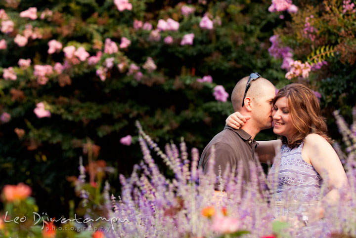 Engaged guy whispers in his fiancée's ear amidst the flower garden. Pre-wedding engagement photo session at Brookside Gardens, Wheaton, Silver Spring, Maryland, by wedding photographer Leo Dj Photography.