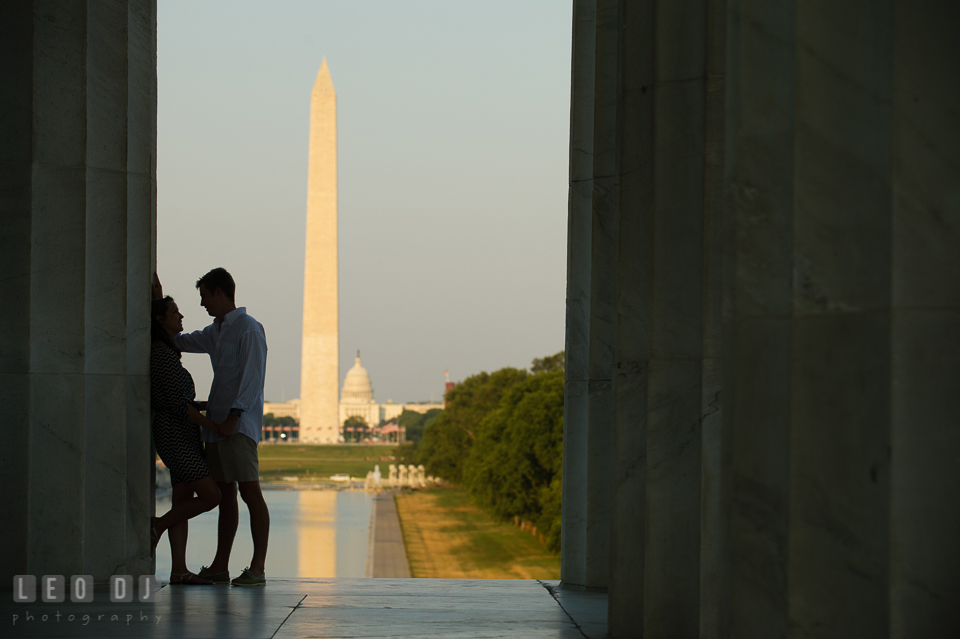 Engaged girl leaning on column, and fiancé about to kiss. Washington Monument, Capitol and reflecting pool in the background. Washington DC pre-wedding engagement photo session at Lincoln Memorial, by wedding photographers of Leo Dj Photography. http://leodjphoto.com