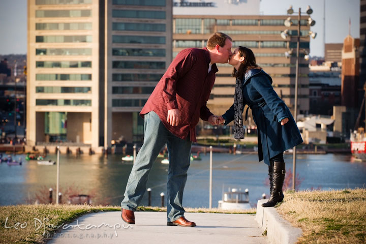 Engaged guy and girl kissing with skyscrapers in the background. Pre-wedding Engagement Photo Session Federal Hill Baltimore and Mother's Grille restaurant bar by wedding photographer Leo Dj Photography