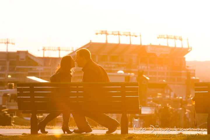 Engaged couple kissing in the sunset light, Ravens stadium in the background. Pre-wedding Engagement Photo Session Federal Hill Baltimore and Mother's Grille restaurant bar by wedding photographer Leo Dj Photography