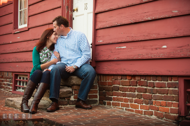 Engaged guy sitting in front of a rustic red house kissing his fiancée. Annapolis Eastern Shore Maryland pre-wedding engagement photo session at downtown, by wedding photographers of Leo Dj Photography. http://leodjphoto.com