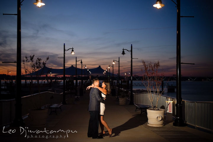 Engaged couple holding close by the pier lights after sunset. National Harbor, Gaylord National Hotel pre-wedding engagement photo session